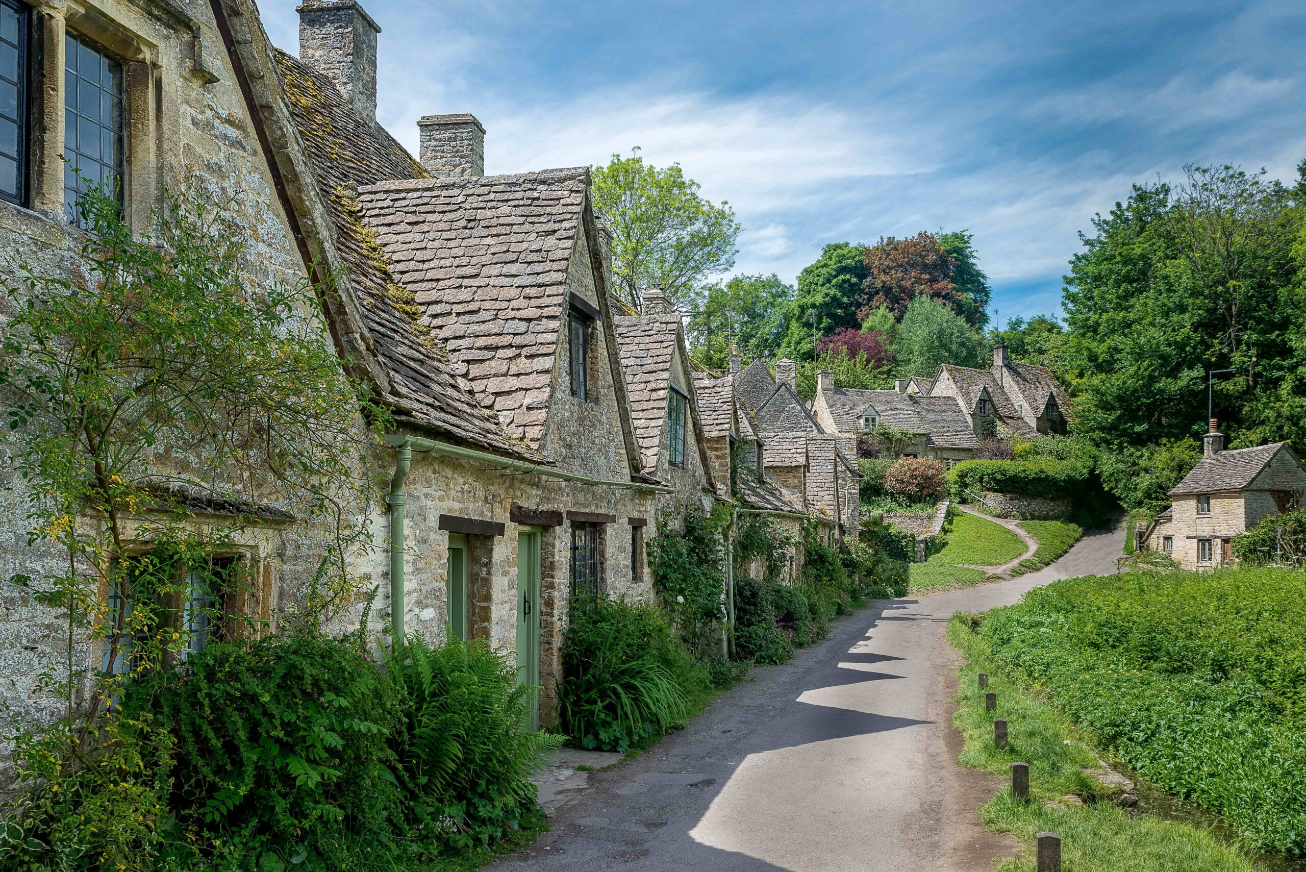 A horizontal view of Arlington Row in Bibury, featuring a row of historic stone cottages set against a lush green backdrop in the Cotswolds.